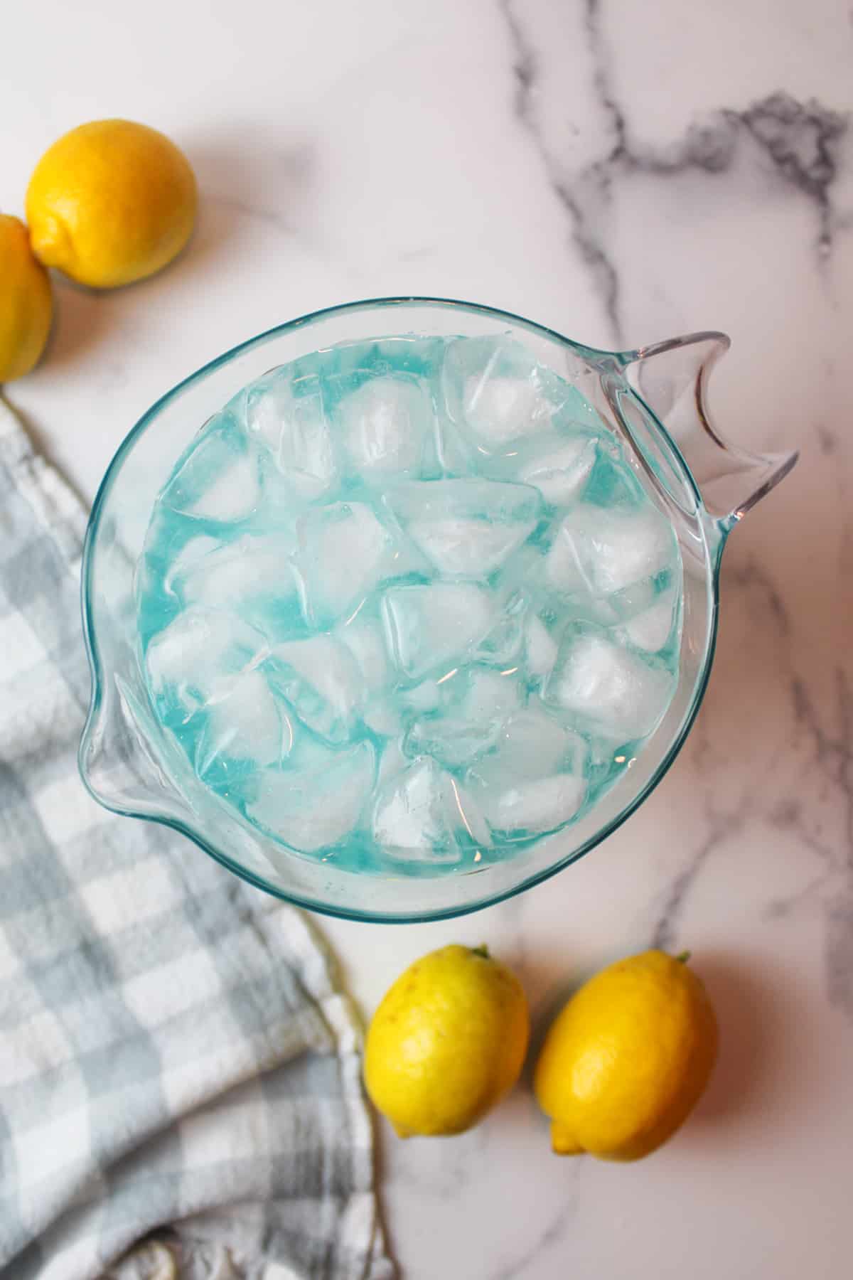 blue raspberry lemonade and ice in a pitcher with fresh lemons to the sides.