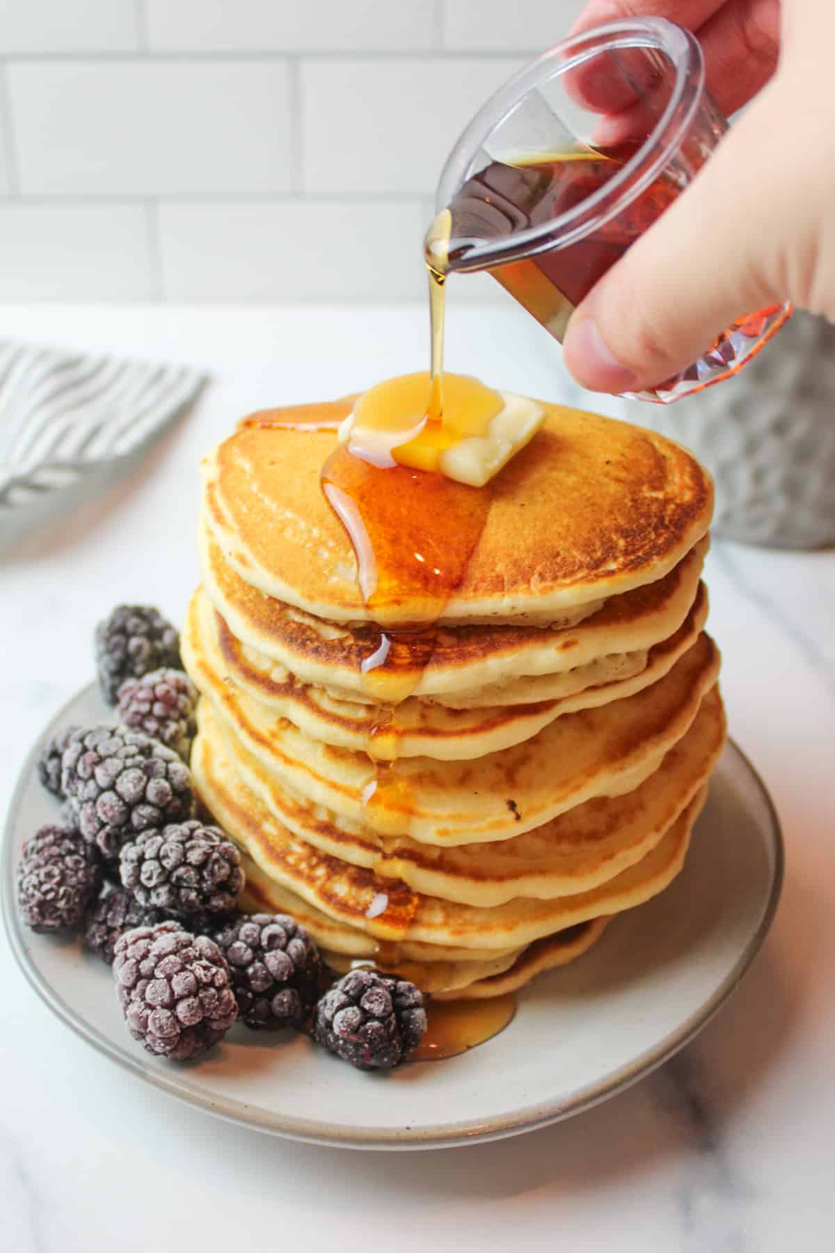 Hand pouring maple syrup over tall stack of golden pancakes, next to frozen blackberries.