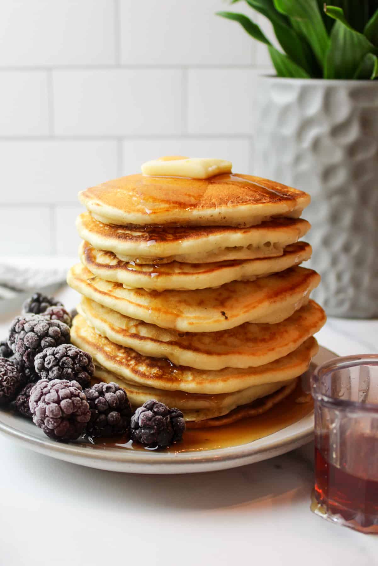 Tall stack of pancakes with butter, syrup, and frozen blackberries on the side.