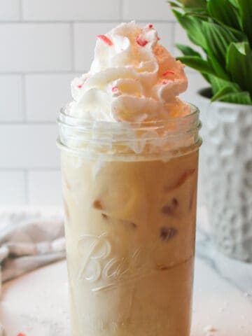 a close up view of side view of a tall glass cup full of peppermint iced coffee topped with whipped cream and peppermint pieces