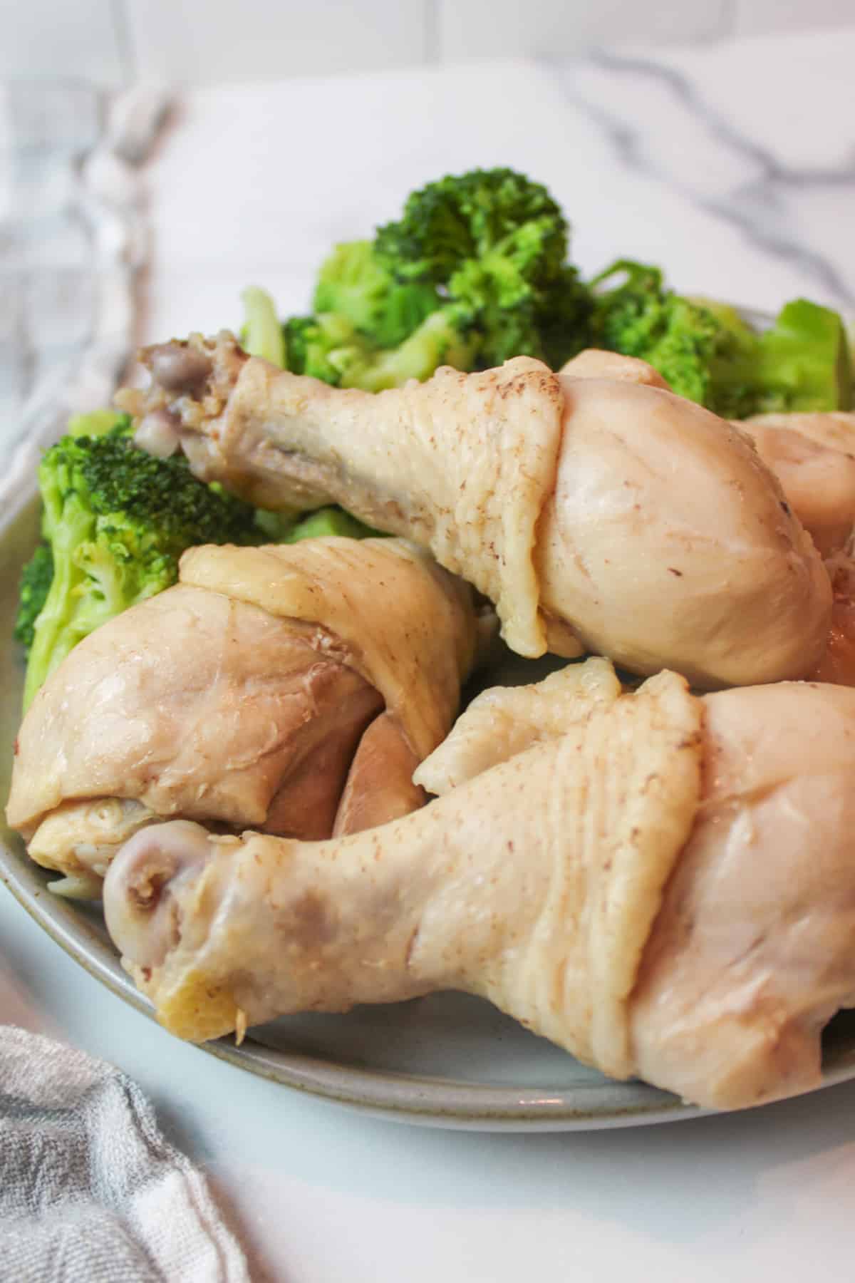 boiled chicken legs piles on a plate with steamed broccoli in the background.