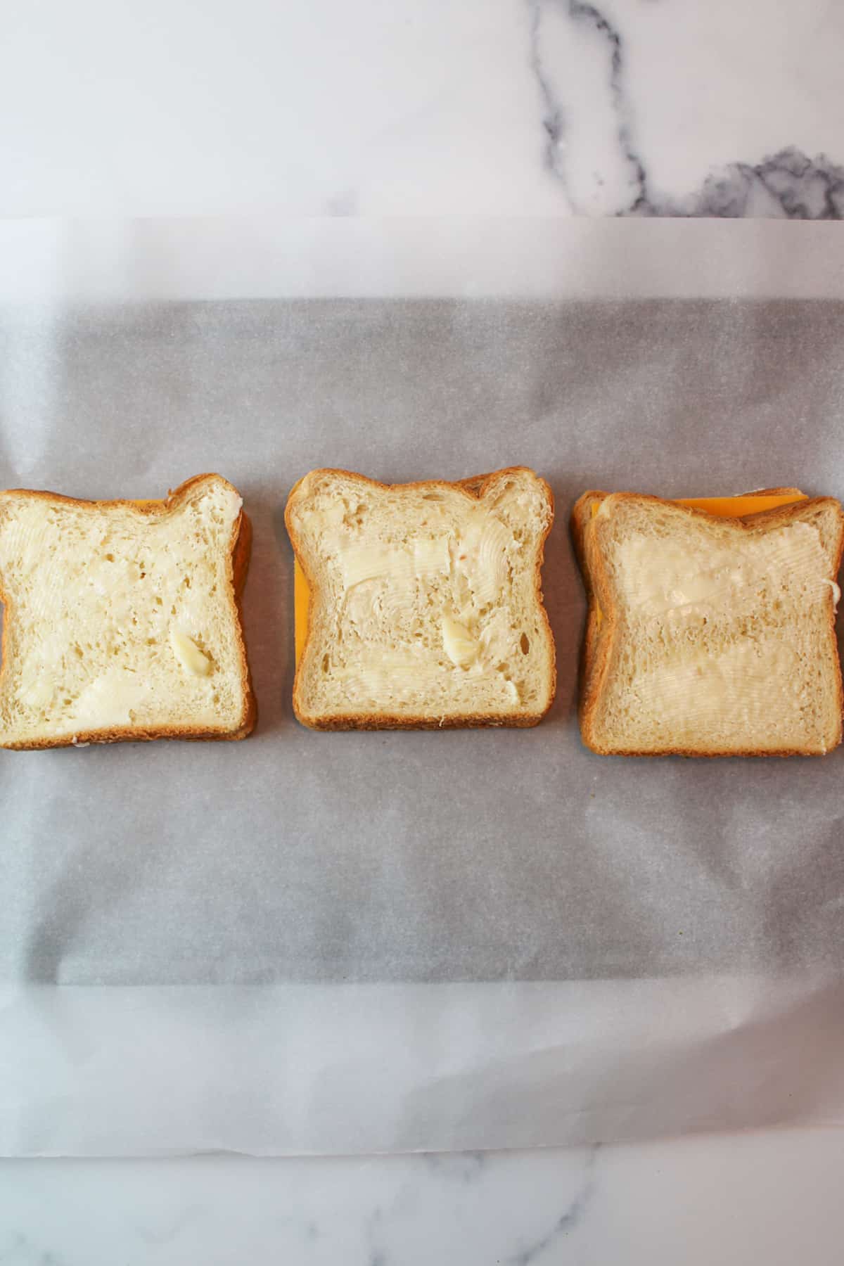 assembled uncooked buttered grilled cheese sandwiches on parchment paper lined baking sheet.