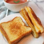 cooked grilled cheeese sandwiches on a plate with a bowl of tomato soup in the background.