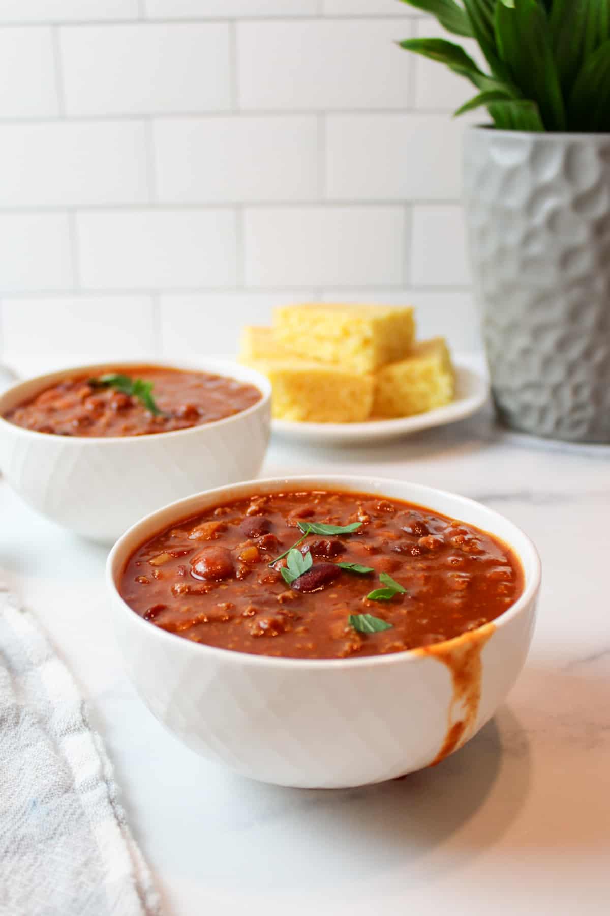 two messy bowls of tomato soup chili with a plate of cornbread in the background.