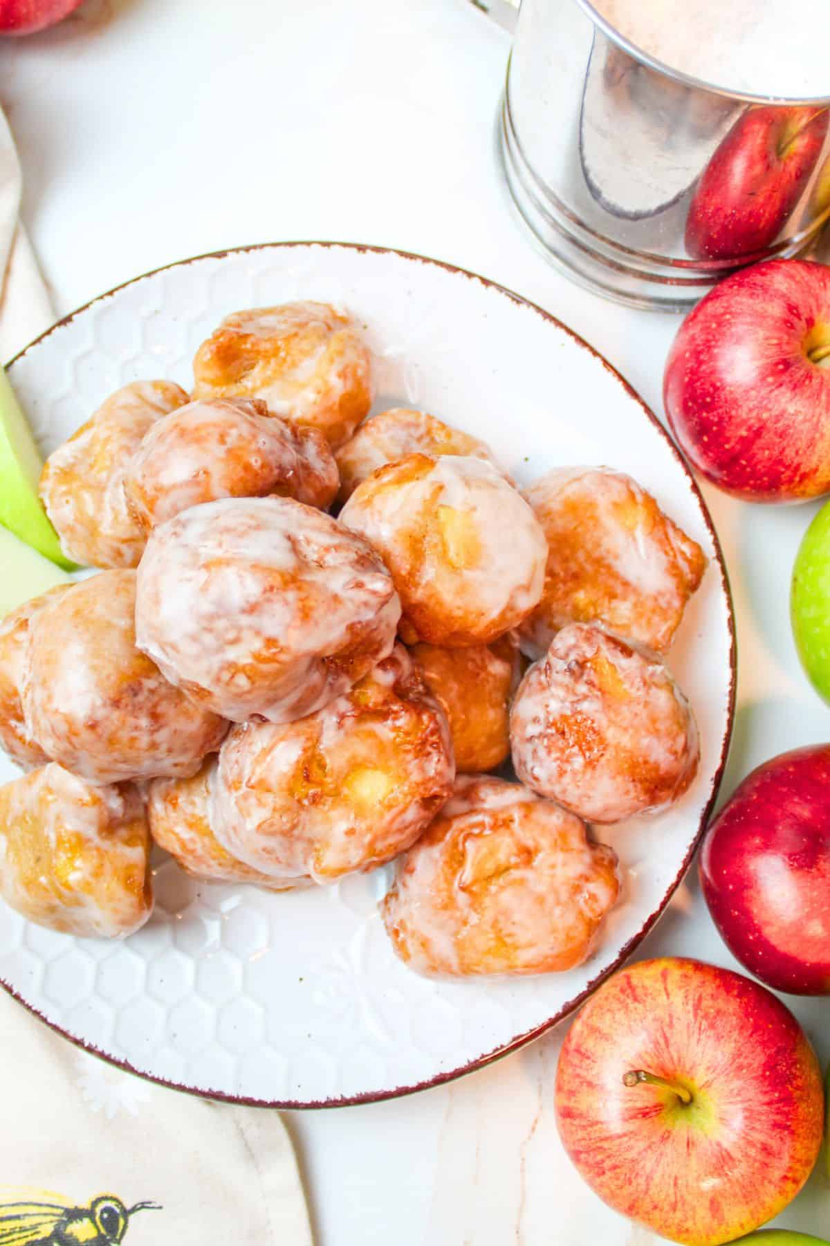 a pile of glazed applesauce fritters on a plate with fresh apples to the side.