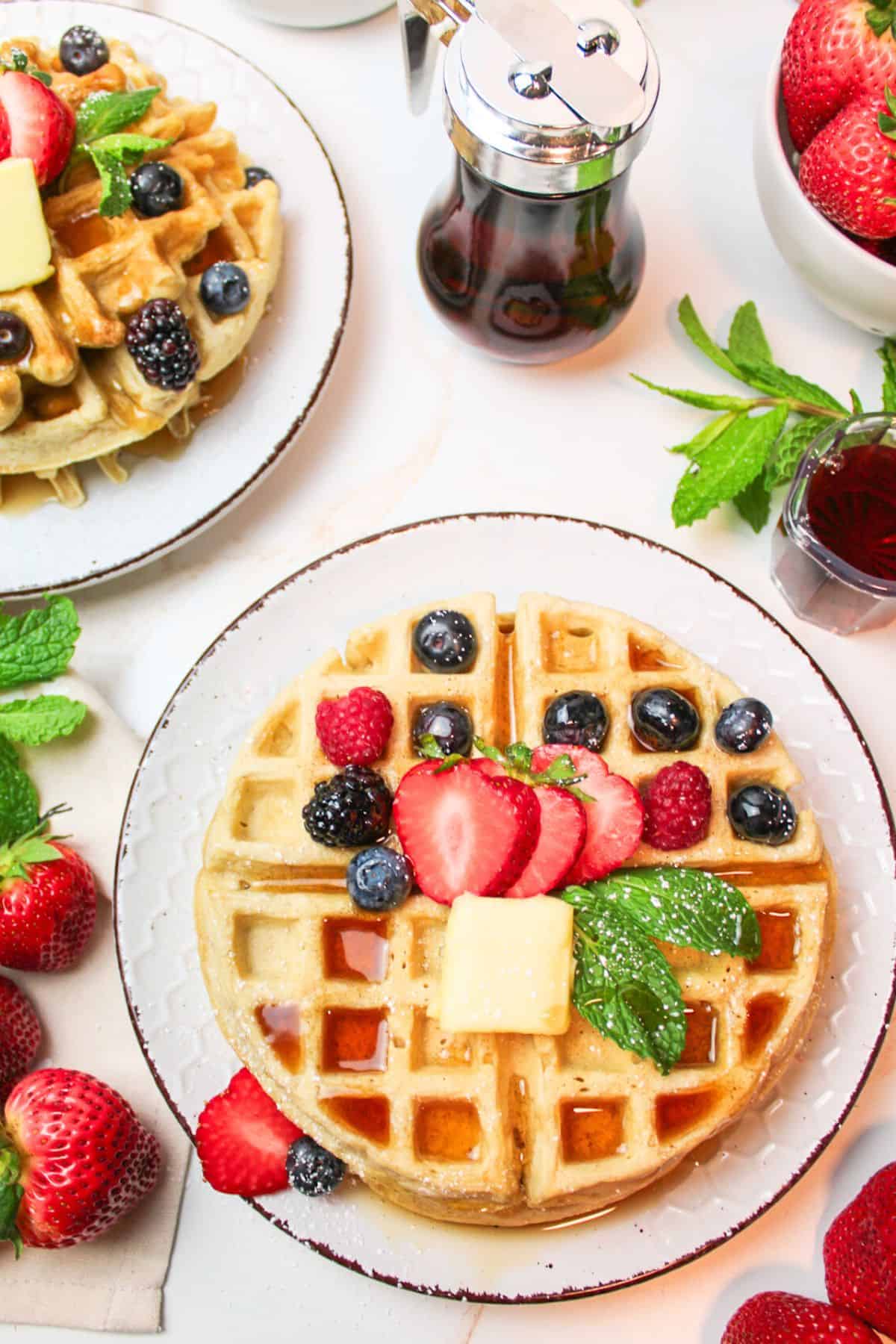 stacked waffles on plates with fresh berries, butter and syrup.