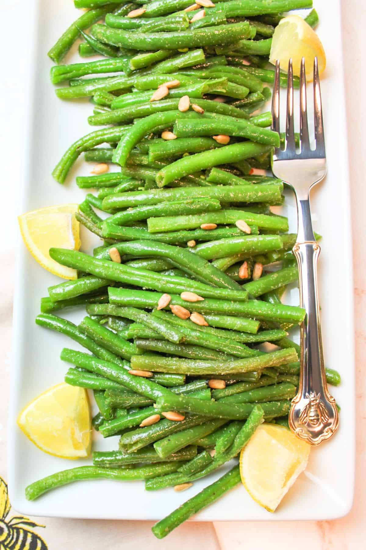 a plateful of cooked green beans with lemon wedges and a large serving fork.