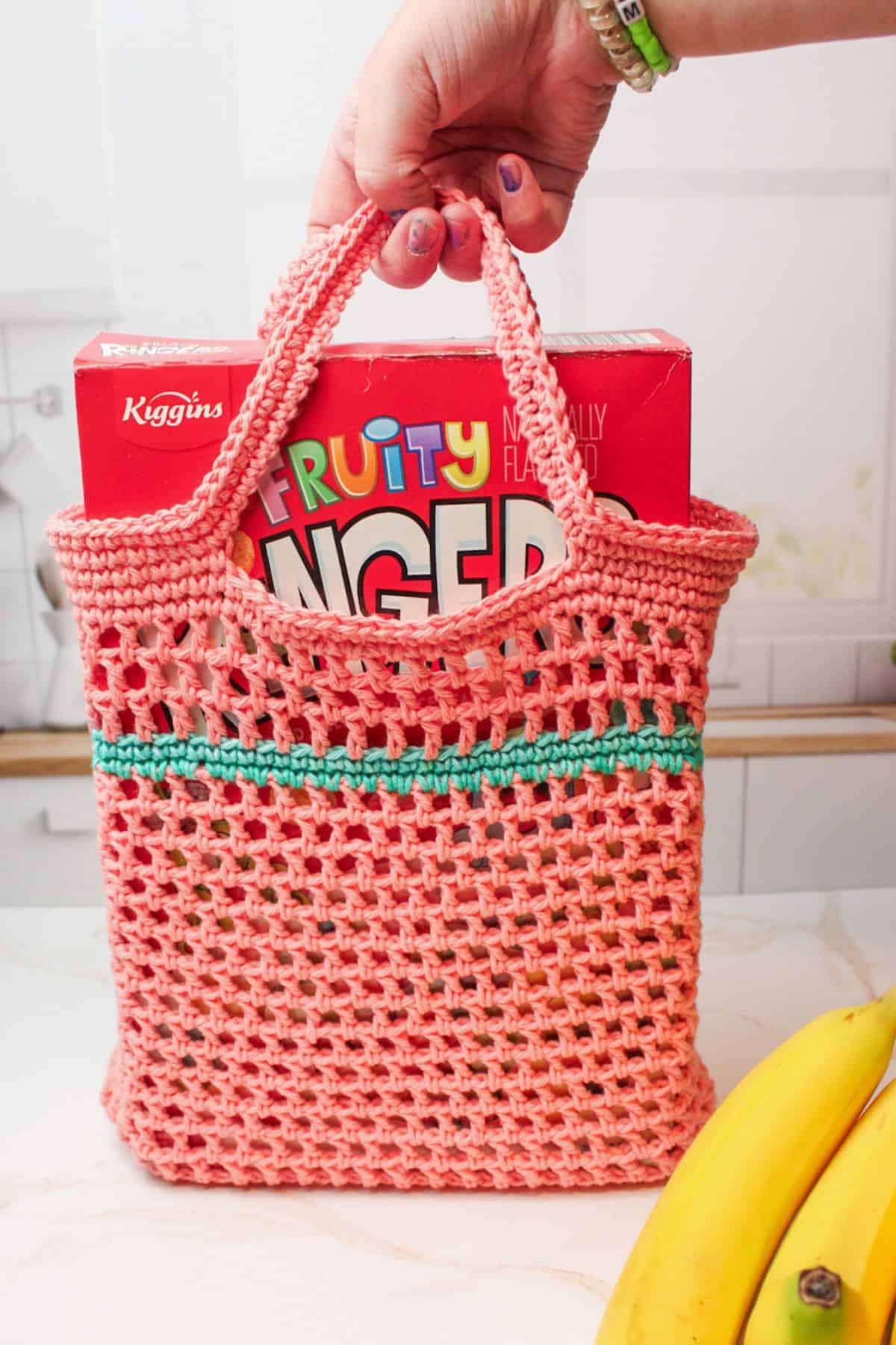 hand pulling up a crochet market bag filled with box of cereal.