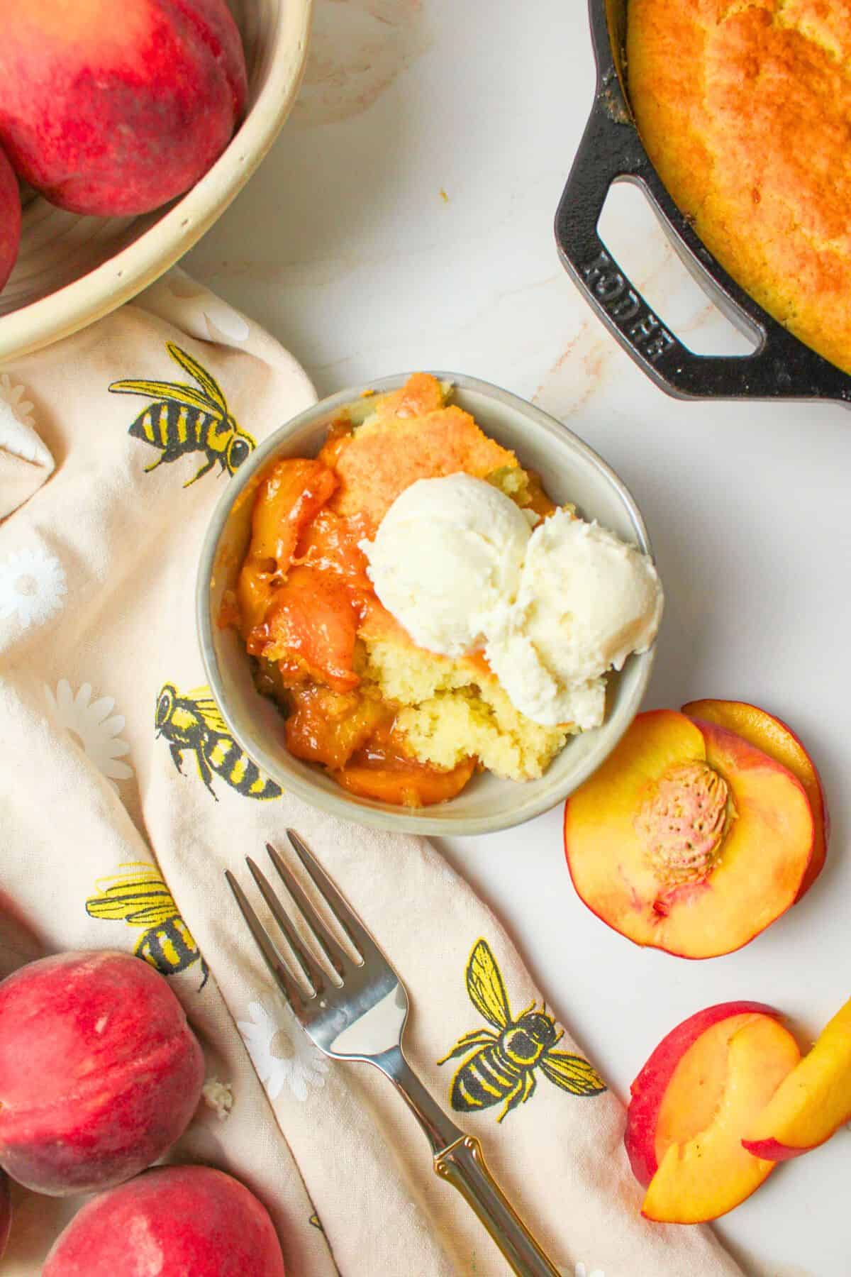 ice cream topped peach cobbler in a bowl with fresh peaches around it.