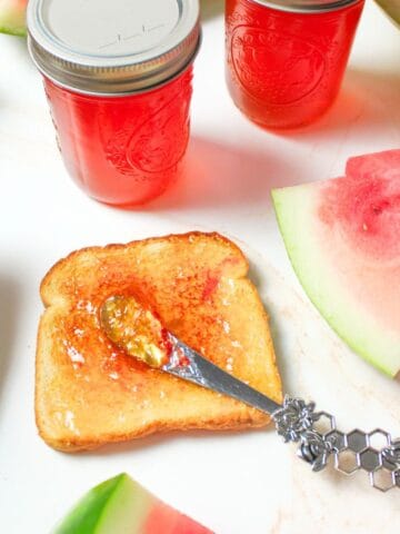 watermelon jelly spread over toast with jars and fresh fruit around it.