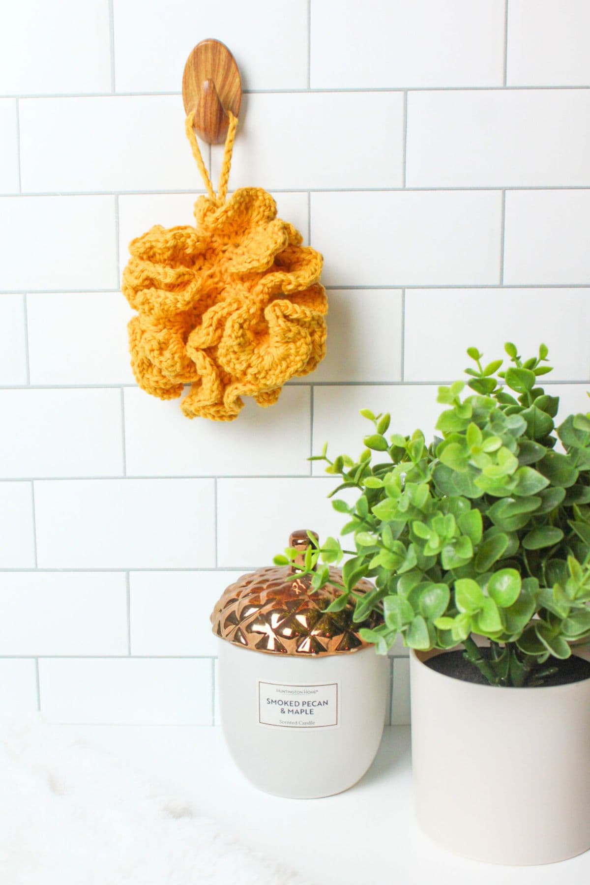 a yellow crochet loofah hanging on a wall by a candle and plant.