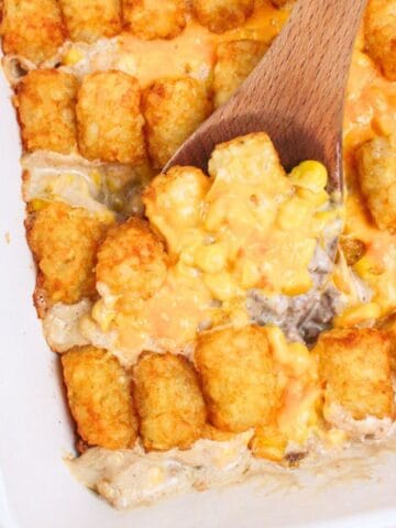 a wooden spoon digging into tater tot corn casserole in baking dish.